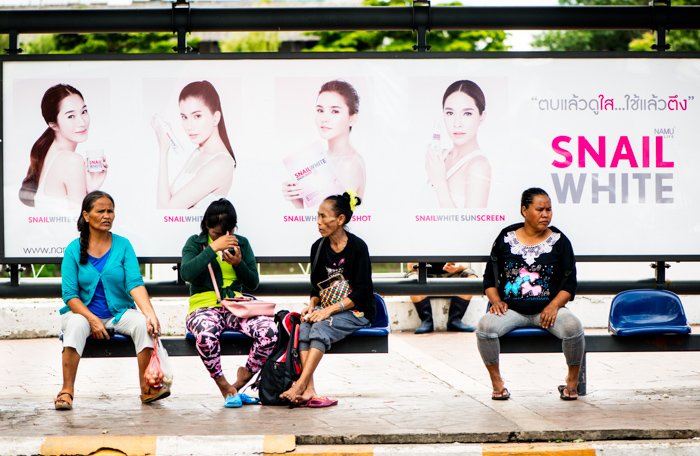 Documentary photography of four Thai women, waiting for a bus, a large advertising billboard behind them