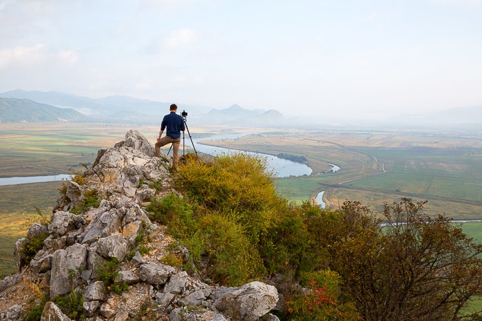 a photographer stands on top a rocky hilltop overlooking a green landscape and river