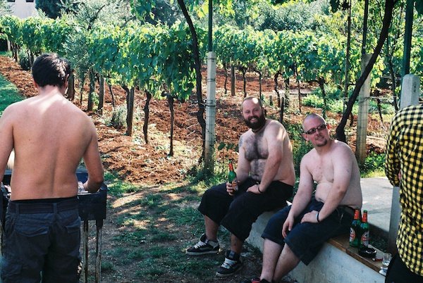 2 over-weight men drinking beer next to a grape vines with another man barbecuing next to them