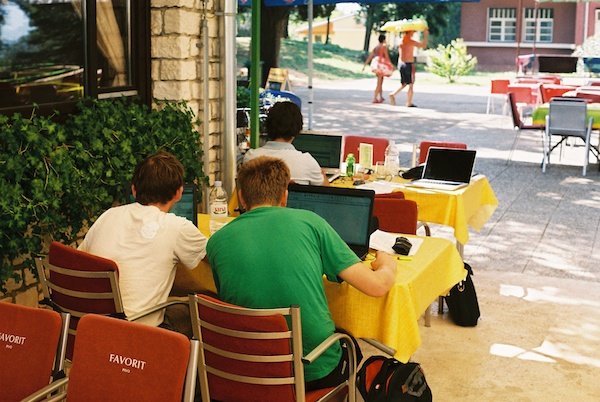 A scene on the terrace of a cafe with 3 young adults with their backs to us working on their laptops
