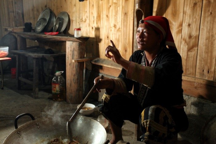Travel photograph in the interior of a lady's home. She is sitting down cooking in a large skillet.