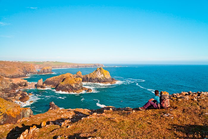 Man sitting at the top of an orange brown rocky mountain, looking at the bright blue sea and sky