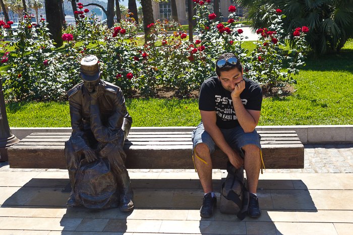 outdoors, in front of a patch of red flowers, a man sits next to a statue, imitating it's thinking posture