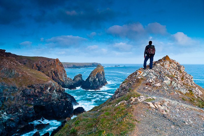 View from behind of a man standing at the top of an outcropping of rock overlooking a rocky shore and bright blue sea and sky