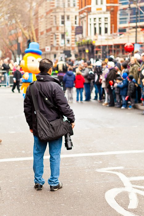 A man with photography gear standing on the street watching the festival crowd in the background